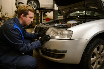 Naklejka na ściany i meble Auto mechanic in car service adjusting headlights on car in the repair shop garage. Auto service, car repair and warranty maintenance concept