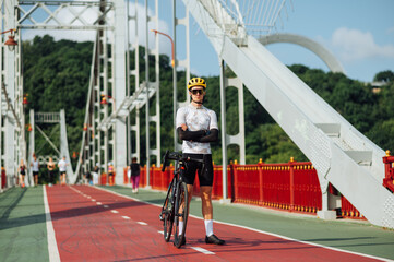 Professional cyclist in sportswear stands with a bicycle on the bridge and poses for the camera with a serious face
