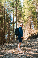 Young male hiker in casual clothes stands in the mountains on a trail through pine trees on a climb and looks back.