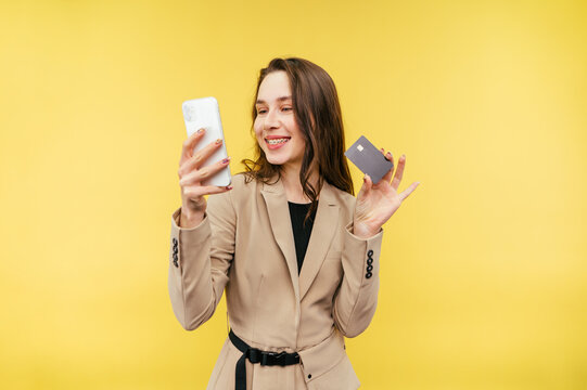 Attractive woman in a suit doing online shopping on a smartphone with a bank card in her hands smiling and looking at the smartphone screen.