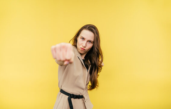 Emotional attractive business woman in a jacket shows a fist to the camera and looks angrily on a yellow background.