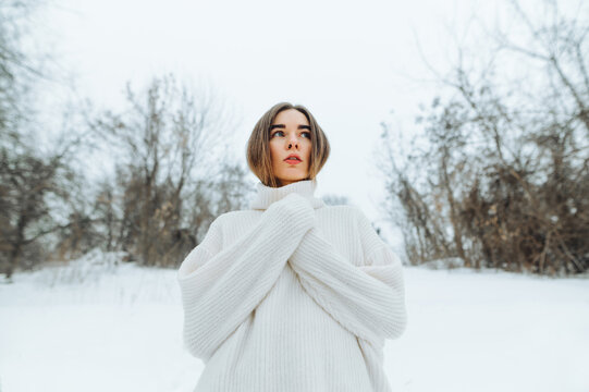 A frozen woman in a poor knitted sweater stands on a snowy winter street and looks away.