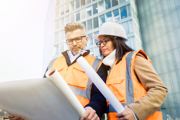 Two engineer colleagues in orange work vests and helmets explore construction documentation. Team...