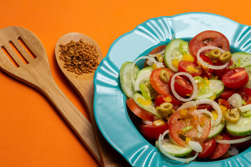 Um prato com salada de vegetais com dois talheres de madeira sobre um fundo alaranjado.