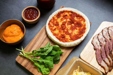 Pizza dough with ingredients on wooden board with black table background. Homemade delicious pizza; top view. 