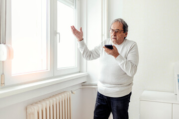 Naklejka na ściany i meble An unhappy elderly man standing next to a cold radiator, using a cell phone to call a technical service. Shot of an angry senior man talks to a technician about a radiator that doesn't work.