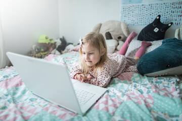 A little girl 5 years old lies on a bed in a children's room and looks into a white laptop and is surprised at what she sees there