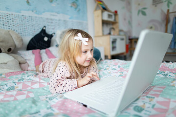 A little girl 5 years old lies on a bed in a children's room and looks into a white laptop and is surprised at what she sees there