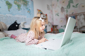 A little girl 5 years old lies on a bed in a children's room and looks into a white laptop and is surprised at what she sees there