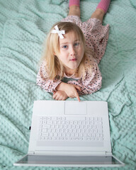 A little girl 5 years old lies on a bed in a children's room and looks into a white laptop and is surprised at what she sees there