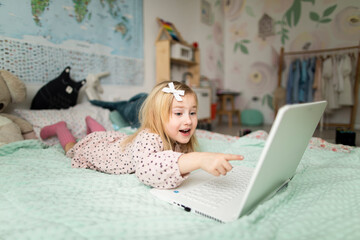 A little girl 5 years old lies on a bed in a children's room and looks into a white laptop and is surprised at what she sees there