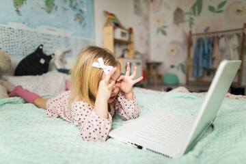 A little girl 5 years old lies on a bed in a children's room and looks into a white laptop and is surprised at what she sees there