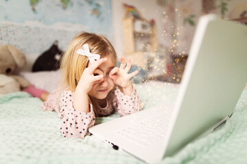 A little girl 5 years old lies on a bed in a children's room and looks into a white laptop and is surprised at what she sees there