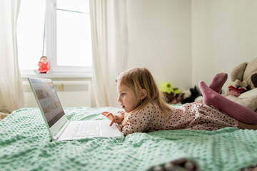 A little girl 5 years old lies on a bed in a children's room and looks into a white laptop and is surprised at what she sees there