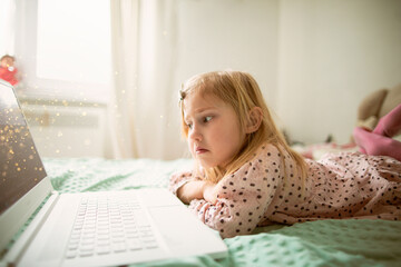 A little girl 5 years old lies on a bed in a children's room and looks into a white laptop and is surprised at what she sees there