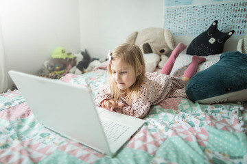 A little girl 5 years old lies on a bed in a children's room and looks into a white laptop and is surprised at what she sees there