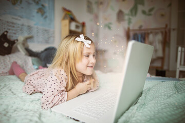 A little girl 5 years old lies on a bed in a children's room and looks into a white laptop and is surprised at what she sees there