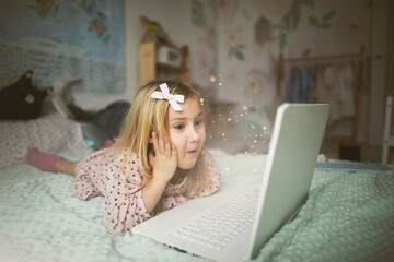 A little girl 5 years old lies on a bed in a children's room and looks into a white laptop and is surprised at what she sees there