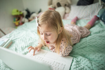 A little girl 5 years old lies on a bed in a children's room and looks into a white laptop and is surprised at what she sees there
