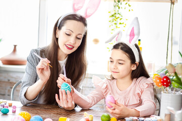Obraz na płótnie Canvas Happy Easter atmosphere! Funny, pretty, creative mom teaching, explain, to her cute, small, joyful daughter how to draw, paint, decorate easter eggs, together wearing bunny ears, sitting at desk