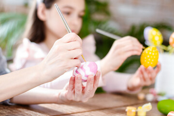 Happy Easter atmosphere! Close up funny, pretty, creative mom teacher, explain, to her cute, small, joyful daughter child how to draw, paint, decorate Easter eggs, together, sitting at desk