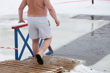 A fat white European believer man goes to the water in an ice hole in the shape of a cross in...
