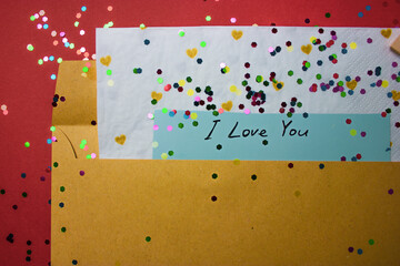 Love letter. A brown envelope with a Valentine's Day message text I LOVE YOU on aa note inside on red terracotta background. Scattered colorful confetti. Festive greeting card for February 14 top view