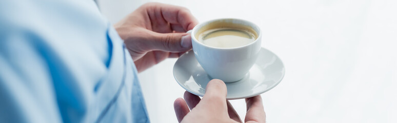 cropped view of man in pajamas holding saucer with cup of coffee, banner.
