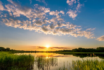 Fototapeta na wymiar Amazing view at scenic landscape on a beautiful lake and colorful sunset with reflection on water surface among green reeds and glow on a background, spring season landscape