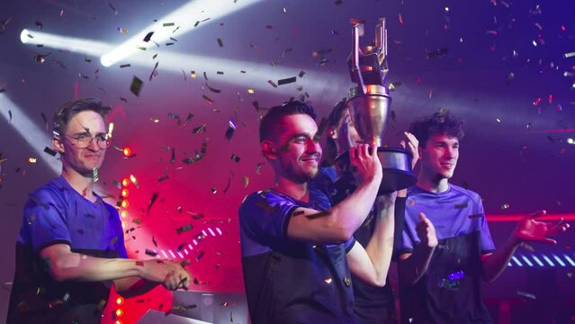 Gamer esportsman with his teammates raise and showing golden cup and waving hand near teammates clapping hands after victory in professional gaming tournament