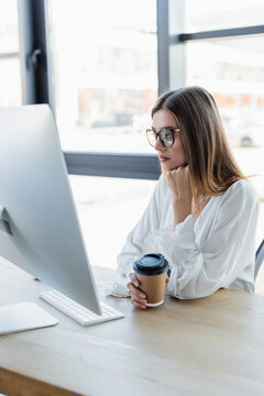 young businesswoman in glasses holding paper cup and looking at computer monitor in office.