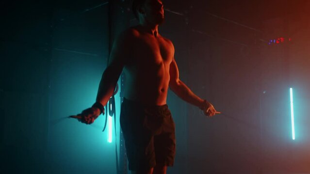 Muscular man with bare torso is jumping using a skipping rope. Male boxer with skipping rope. Fighter doing jump rope exercises in colorful light with smoke in slow motion.