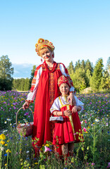 A young blonde woman and a Caucasian child girl in a red national dress with a red shawl and a kokoshnik. A woman stands on a field with flowers and hugs a child.