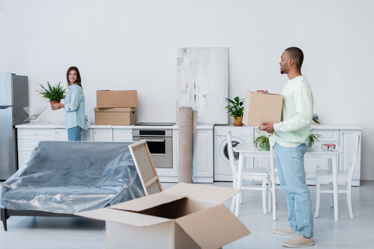 happy african american man holding carton box while looking at girlfriend with plant during relocation.
