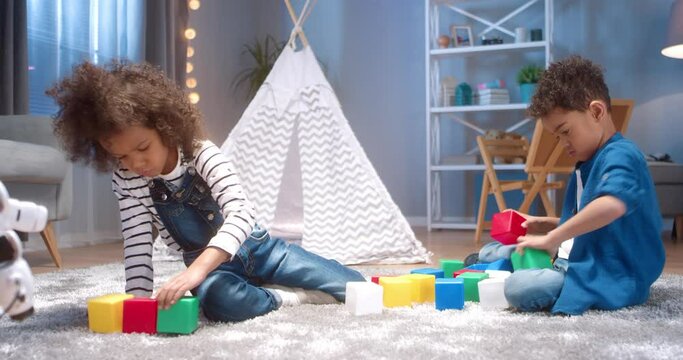 Cute little African-American brother and sister play home sitting on floor and share corstructure to build separately.