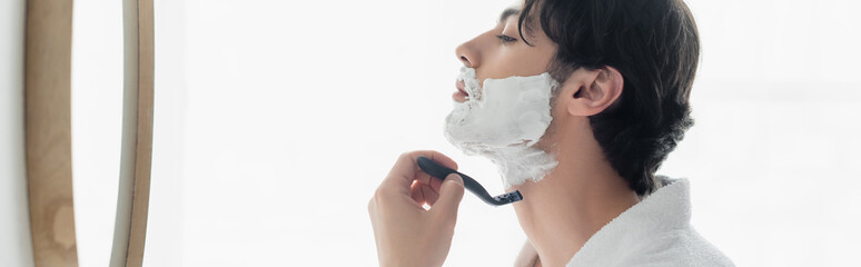 side view of young brunette man shaving near mirror in bathroom, banner.