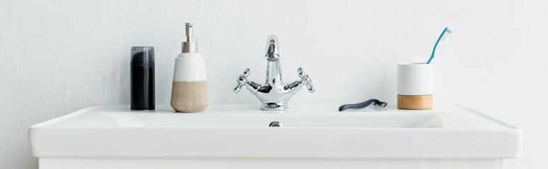 toothbrush, safety razor, liquid soap and shaving foam on white sink with faucet, banner.