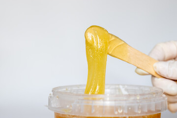 woman in white glove is holding a wooden spatula with sugar or wax paste for depilation. grey background, beauty concept. space for text. plastic jar with sugar pasta.