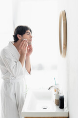 side view of man in white bathrobe cleaning face with cotton pad in bathroom.