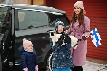 Kids with cat on hands against car in the yard of house at winter. Mother hold flag of Finland.