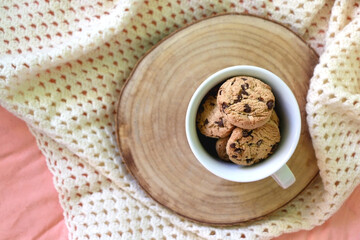 Fototapeta na wymiar Mug filled with chocolate chip cookies and soft knitted blanket on a bed. Flat lay.
