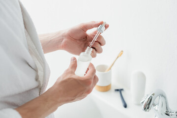 cropped view of man holding bottle with cosmetic serum in bathroom.