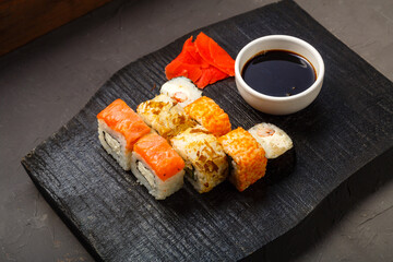 Assorted salmon and shrimp rolls on a black board next to soy sauce and ginger on a dark background.