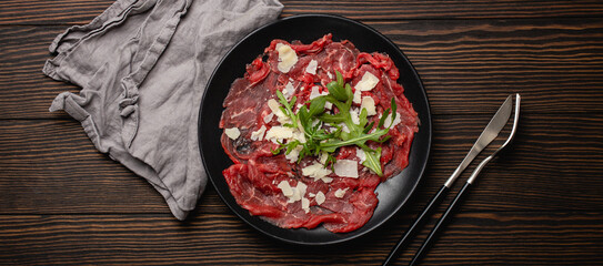 Italian cold meat appetizer Beef carpaccio with parmesan cheese and arugula on black plate with fork and knife on dark brown wooden rustic background flat lay from above 
