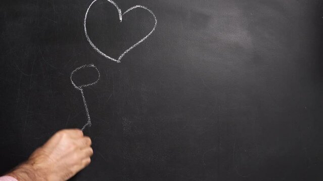 A man's hand in a red and white striped shirt draws a man with a heart on a string on the blackboard, giving it to another man. A holiday concept for Valentine's Day. February 14