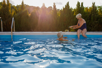 Senior woman pouring lemonade into two glasses while sitting on the poolside and her husband swimming - Powered by Adobe
