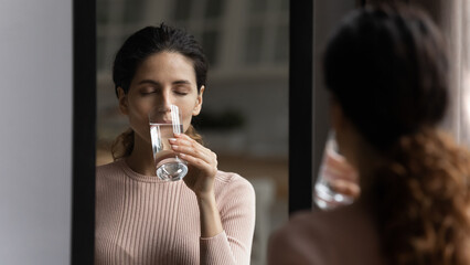Young attractive woman reflecting in mirror while standing indoor alone holding glass and drinks still water, close eyes enjoy fresh beverage, reduces thirst. Healthy life habit, refreshment concept