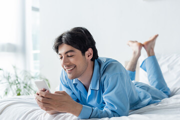 cheerful barefoot man in blue pajamas messaging on mobile phone in bedroom.
