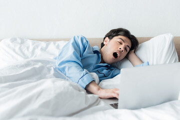 sleepy man yawning near laptop while lying in bed in morning.