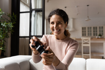 Cheerful Latina woman gamer hold gamepad controller play videogames sit alone on couch in living room. Console online video games lover, home hobby, weekend activities, modern tech amusements concept